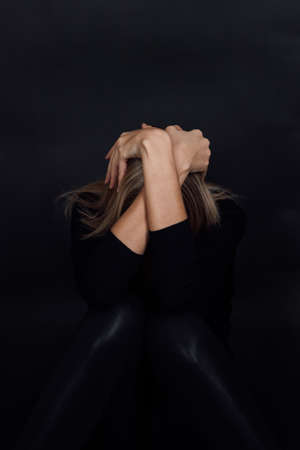 Blonde woman in black dress hiding face with arms dressed in black clothes in depression with black background. Mental and physical abuse. Rough patchの写真素材