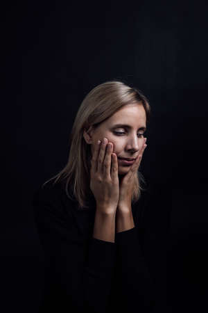 Pretty depressed woman with hands on cheeks in black dress on black background. Victim of physical...