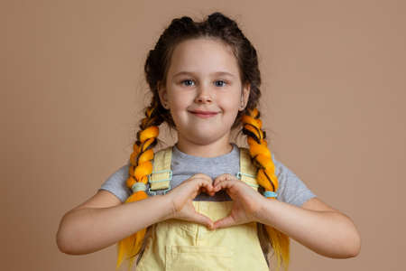 Kind little smiling girl, with yellow kanekalon pigtails looking at camera showing heart with...