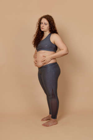 Vertical image of fat excess weight upset barefoot disappointed woman, holding tummy. Oversize...