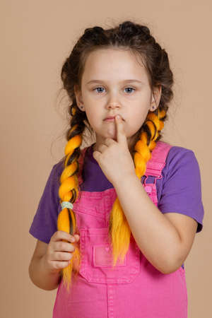 Pensive small, female child with serious eyes looking at camera with yellow kanekalon braids touching mouth with one finger in pink jumpsuit and purple t-shirt on beige backgroundの写真素材