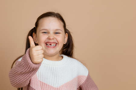 Portrait of young beautiful joyful smiling girl with long dark hair in white, pink sweater standing, showing thumbs up sign, gesture of approval on brown background.の写真素材