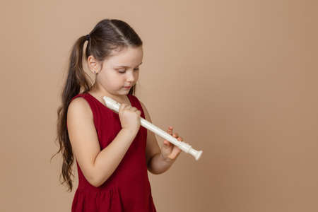Girl in red dress hold common flute with head down and clamp holes on it, beige background. Learning to play woodwind musical instrument.の写真素材