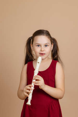 Girl in red dress thoughtfully hold common flute and pinch holes on it, beige background. Learning to play woodwind musical instrument.の写真素材