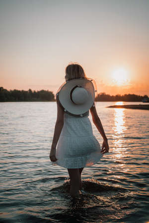 Young woman on the background of the sunset rear view. Photo of beautiful blonde in a white dress and a straw hat around her neck walking in the water and holding the hem of the dress with her handsの写真素材