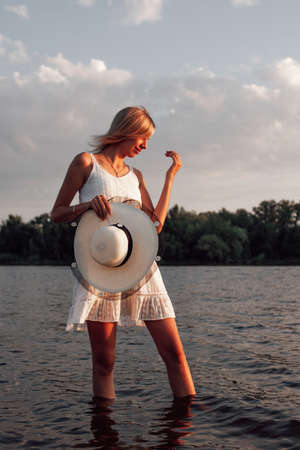 A woman is standing in the water. A beautiful young blonde in a white summer dress and a white straw hat with a black ribbon in her hand poses in the water against the background of the landscapeの写真素材