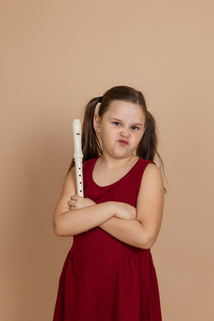 Girl in red dress with displeased expression hold flute with arms crossed, beige background. Learning to play woodwind musical instrument.の写真素材