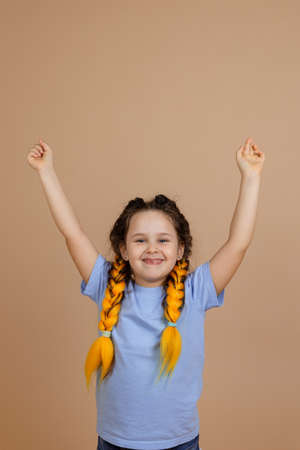 Happy young girl smiling celebrating with hands up looking at camera with yellow kanekalon braids on head on beige background wearing blue t-shirt and jeansの写真素材