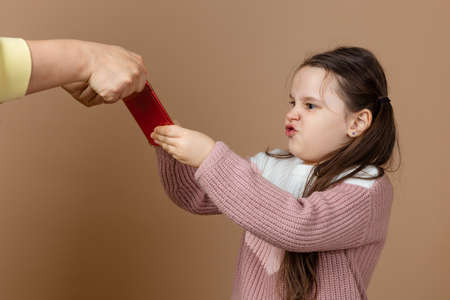 Portrait of parent taking smartphone out from girls hands, beige background. Discontent daughter hold tight phone and oppose. Concept of prevention of telephone addiction in children, nomophobiaの写真素材