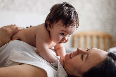 Closeup portrait of happy young mother and baby lying on bed in room. Carefully hold naked baby in arms on chest. Selective focus, free copy space. Concept of maternal affection and childcareの写真素材