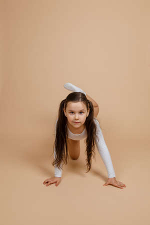 Portrait of young cute concentrated girl with long dark hair in white training swimsuit and socks standing on all fours raising leg up, posing on brown background.の写真素材
