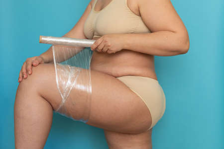 Woman in beige underwear wrap thigh with cling film closeup side view. Body wrapping for weight...