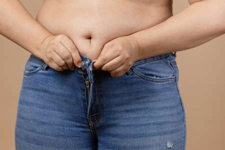 Caucasian female with big belly and overweighted sides trying to zip up blue jeans. Visceral fat. Body positive. Sudden weight gain. Tight little clothes. Need for wardrobe changeの写真素材