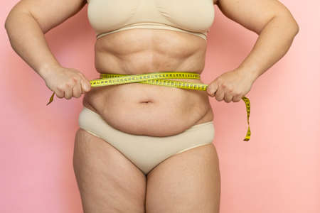 Cropped photo naked after birth woman tummy, measuring her waistline by tape. Fat abdomen and sagging folds, excess skinの写真素材