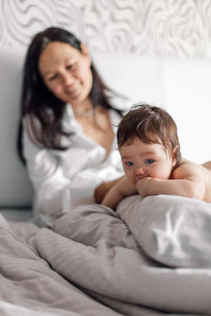 Closeup portrait of naked baby lying on tummy on gray bed, free copy space. Happy mother on blurred background watching her little daughter in bedroom. Concept of maternal affection and childcareの写真素材