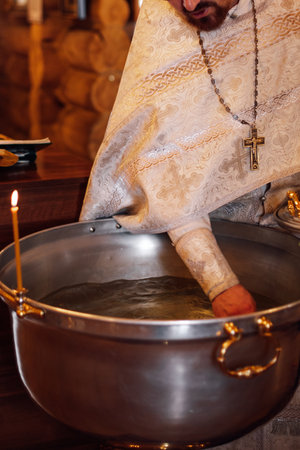 Unrecognizable bearded priest in silver cassock stand at metal font, consecrating water for ceremony...