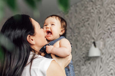 Portrait of young mother with little daughter in bedroom closeup, free copy space, blurred background and green plants. Hold in hand screaming baby. Concept of maternal affection and childcareの写真素材