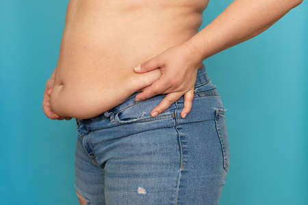 Side view of unrecognizable fat plump woman standing in blue jeans, squeezing excess naked waist on...