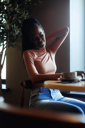 Young afro american woman stretching neck with closed eyes sitting on table and working in coffee house, side view, blurred foreground. Black girl working remotely, freelancer. Sore neck, tirednessの写真素材