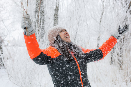 Joyful middle aged woman with widely outstretched arms looking up at snow falling down on her on walk in snowy forest. Magic winter time full of white colorの写真素材