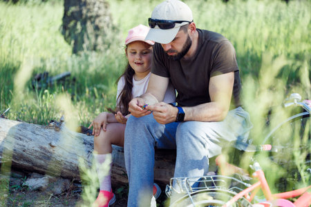 Portrait of family wearing caps, sitting on dry log in park. Little cute girl daughter looking at hands of man father.の写真素材