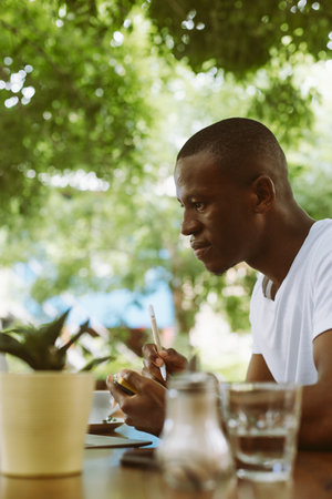 Vertical concentrated modern dark skin man in casual clothes writing business plan in textbook during webinar on laptopの写真素材