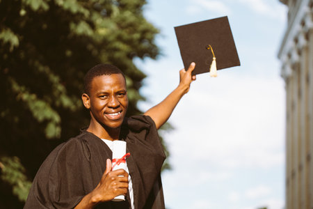 Joyful african american graduate from university stand outdoors in mantle and holding in hands...