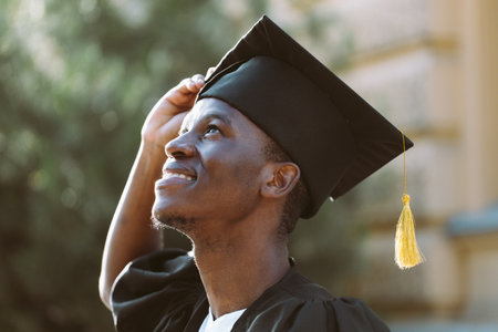Portrait closeup of afro american student in graduation mantle and hat standing outdoors and with pleasure looking up. Job search, graduate from university, start in life, thoughts about futureの写真素材