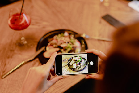 Cropped photo of woman sitting near salad and glass with red cocktail, holding smartphone, taking pictures of food.の写真素材
