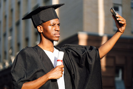 Serious african american graduate from university taking photo with front camera of phone with higher education diploma. Student in black mantle and hat. Future career, graduation, young specialistの写真素材