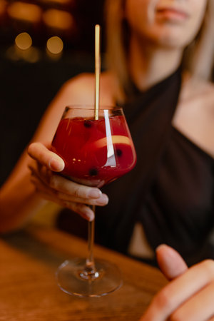 Cropped photo of young woman sitting at table in restaurant stretching hand, holding glass with red cocktail with straw.の写真素材