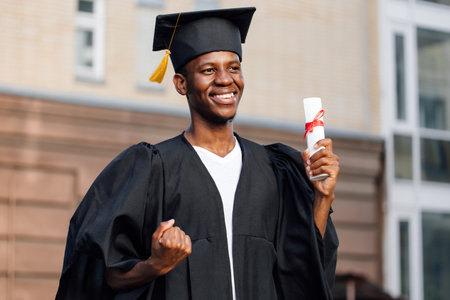 Portrait of happy african american graduate from university standing outdoors near building and holding in hand higher education diploma, selective focus. Student, graduation, ceremonial celebrationの写真素材