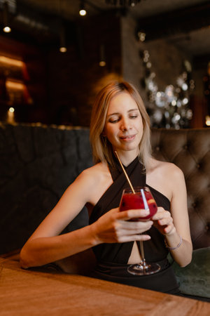 Portrait of young happy good-looking woman sitting at table in restaurant, holding glass with red cocktail with straw.の写真素材