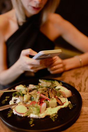 Cropped photo of young woman sitting on sofa near black plate with vegetable salad, holding smartphone in restaurant.の写真素材