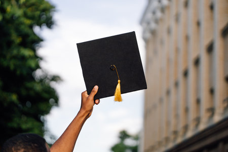 Photo back view of african american graduate from university standing outdoors and holding in hand black hat, blurred sky background, free copy space. Future career, graduation, young specialistの写真素材