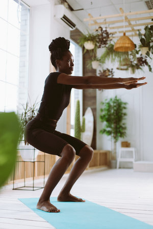 Athletic afro american woman squatting with arms extended forward side view, training on gymnastic...