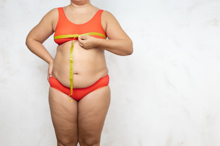Overweight woman measure chest circumference with tape, white background. Naked woman in red underwear with large hanging belly and cellulite body. Weight loss, loose skin, plus size peopleの写真素材