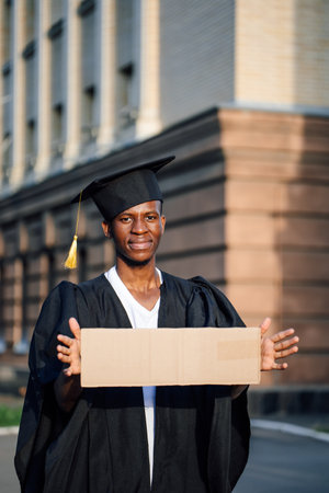 Poor black guy standing with cardboard sign on street looking for job. University or college graduating student wearing graduate gown and cap. Education, career, employment problem, vacancyの写真素材
