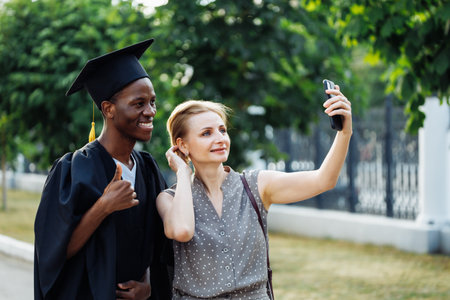 Cheerful afro american student in black graduation mantle and hat with attractive woman take selfie on smartphone. Job search, graduate from university, like, finger up, ceremonial celebrationの写真素材