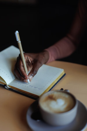 Cropped photo of African-American woman sitting at table with cup of coffee, glasses in cafe, taking notes in notebook.の写真素材