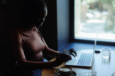 Side view of young busy African-American woman sitting at table in cafe, typing working on laptop, giving credit card.の写真素材
