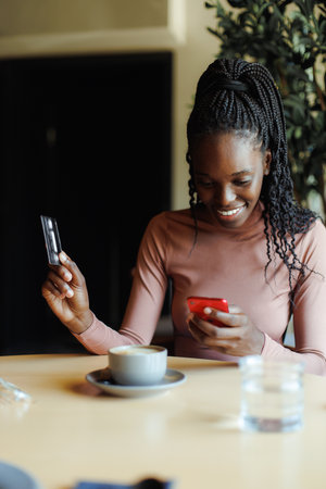 Portrait of young focused busy African-American woman sitting at table in cafe using red smartphone, giving credit card.の写真素材