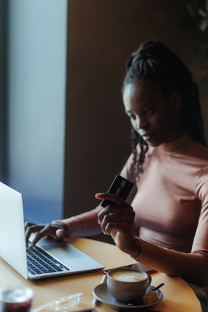 Portrait of young smiling cute African-American woman sitting in cafe, typing working on laptop, holding credit card.の写真素材