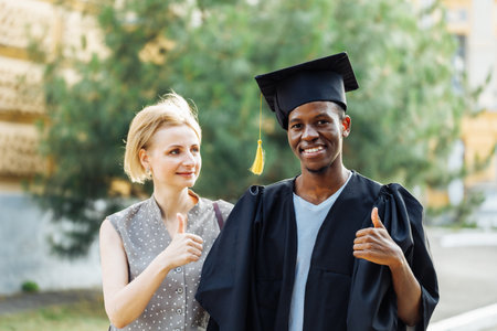 Portrait of afro american student in graduation mantle and hat standing outdoors with happy middle aged woman. Higher education, graduate from university, like, finger up, ceremonial celebrationの写真素材