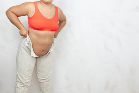 Hands of overweight woman massage hanging folds on belly, white background. Woman in red underwear stand with unbuttoned white jeans. Overeating, fat body, plus size people, body positiveの写真素材