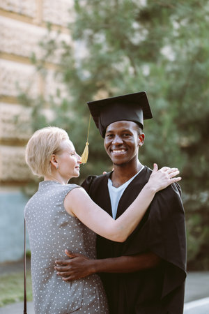 Joyful afro american student in graduation mantle and hat gratefully hugging middle aged woman....