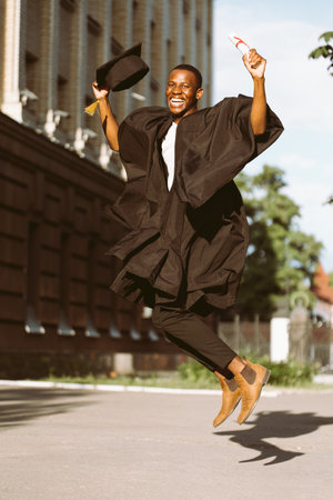 Happy black guy graduate from university with pleasure jumping up in black mantle and hat. Photo of student outdoors on sunny day. Higher education diploma, future career, graduation, celebrationの写真素材