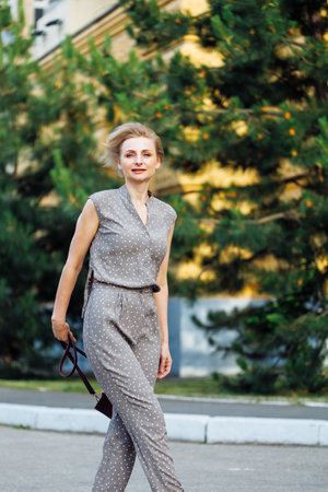 Portrait side view of 40 years old woman walking outdoors among green firs with burgundy leather shoulder bag in hand. Middle aged lady in nice costume. Selective focus, blurred urban backgroundの写真素材