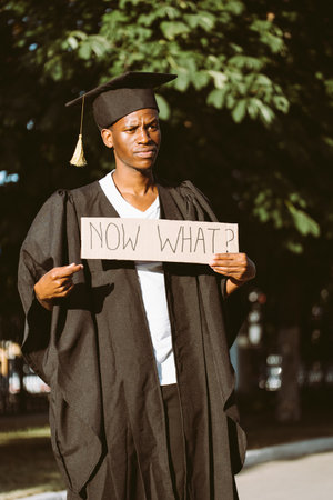Portrait of disappointed afro american guy holding cardboard poster and lending hand on street. Looking for job, hiring and employment issue. University or college graduating student in gown and capの写真素材