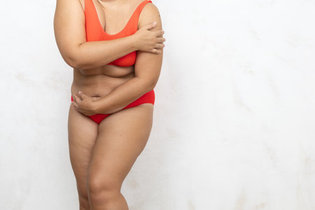 Hands of overweight woman cover naked body, white background. Woman in red underwear with cellulite hips and large belly embarrassed body. Overeating, fat figure, plus size peopleの写真素材